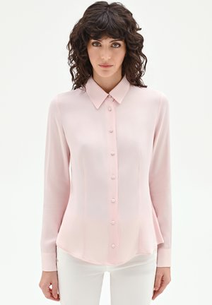 Donna con capelli ricci e scuri che indossa una camicetta rosa chiaro a maniche lunghe con bottoni e pantaloni bianchi, in piedi davanti a uno sfondo bianco semplice.