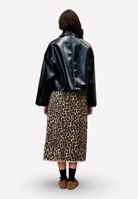 Vrouw die wegkijkt, gekleed in een zwart leren cropped jack, een midi-rok met luipaardprint, zwarte sokken en bruine fuzzy pantoffels.