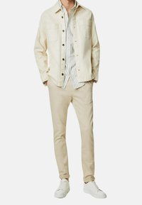 Chaqueta de utilitario de algodón beige con dos bolsillos en el pecho, camisa de rayas blanca y azul por debajo, combinada con pantalones beige claros y zapatillas blancas.