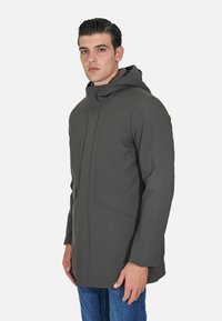 Uomo con giacca con cappuccio grigio scuro e tasche frontali, in piedi leggermente di lato contro uno sfondo chiaro e uniforme.