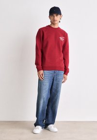 Felpa rossa con un piccolo logo bianco, abbinata a jeans blu larghi e scarpe da ginnastica bianche. Il modello indossa un cappellino blu navy.