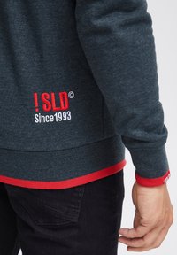 Sweat-shirt gris foncé avec accents rouges. Présente un texte brodé "! SLD" et "Since 1993" à l'ourlet. Poignets et taille côtelés visibles.