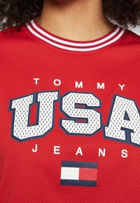 Jersey de malla rojo con el atrevido texto en blanco "USA" y "TOMMY JEANS". Cuello de tripa a rayas y un pequeño logo gráfico de bandera en rojo, blanco y azul.