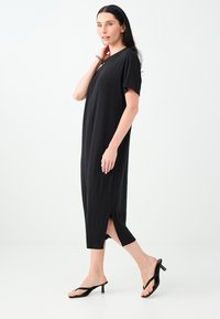 Femme aux longs cheveux foncés porte une robe midi noire ample avec une fente sur le côté et des sandales noires à talons, se tenant devant un fond blanc uni.