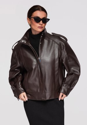 Kobieta w ciemnobrązowej, fakturowanej skórzanej kurtce, czarnej sukience z golfem, okularach przeciwsłonecznych w kocim stylu oraz złotych kolczykach kołach na jasnym tle.