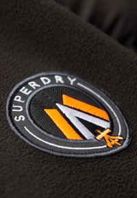 Sort fleece stof har et cirkulært broderetmærke med "SUPERDRY" tekst, et stiliseret "M" i orange og hvid, samt et snefnugsikon.