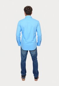 Camisa de manga larga azul clara con dobladillo curvado y puños abotonados, combinada con jeans azul oscuro y zapatos marrones. Vista desde atrás.