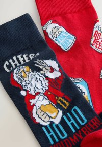Rote und marineblaue Socken mit einem Santa, der Bier hält, im Design, Text "Prost" und "Ho Ho Halt mein Bier" sowie gestrickten Mustern in lebendigen Farben.