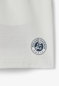 Camiseta atlética blanca con un patrón de cuadrícula, que presenta un parche con el logo azul "Roland Garros París" cerca del dobladillo.