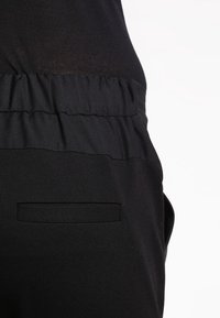 Svart tyg med struktur som visar en sammanknuten midjeband och en platt fickdetalj, vilket betonar en stilren design och monokrom färg.