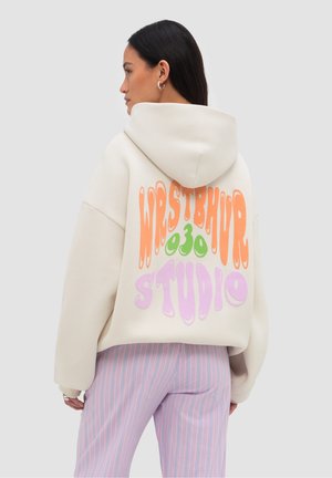 Kobieta nosząca oversize kremową bluzę z kapturem z pomarańczowym, zielonym i lawendowym napisem "WRSTBHVR 030 STUDIO", w połączeniu z różowymi spodniami w paski.