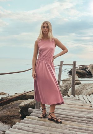 Donna con vestito rosa senza maniche e sandali neri in piedi su un molo di legno vicino a una costa rocciosa sotto un cielo nuvoloso.