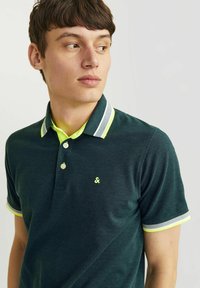 Polo shirt vert foncé avec une patte de boutonnage, présentant des accents jaune fluo sur le col et les manches, ainsi qu'un petit logo sur la poitrine.