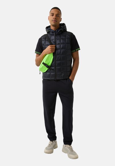 Schwarze Pufferweste mit Kapuze, grüne Akzente auf einem schwarzen Shirt, marineblaue Hose und hellgraue Sneakers. Enthält eine kleine neongrüne Tasche.