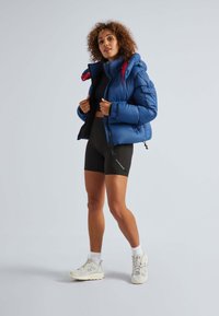 Blauwe pufferjas met rode accenten, voorritssluiting en zijzakken, gedragen over een zwarte crop top en shorts, gecombineerd met witte sneakers.