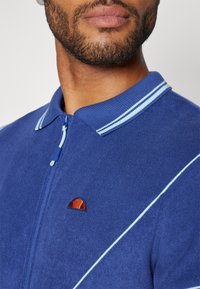 Blå glidelås cardigan laget av teksturert stoff med en kontrasterende lyseblå krage og stripete detaljer, med en liten logo på brystet.
