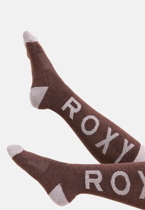 Ruskeat sukat, joissa on valkoiset varvas- ja kantapääkorostukset, ja joissa on suuri "ROXY" -painatus sivuilla, valmistettu tekstuurisesta neulomateriaalista.