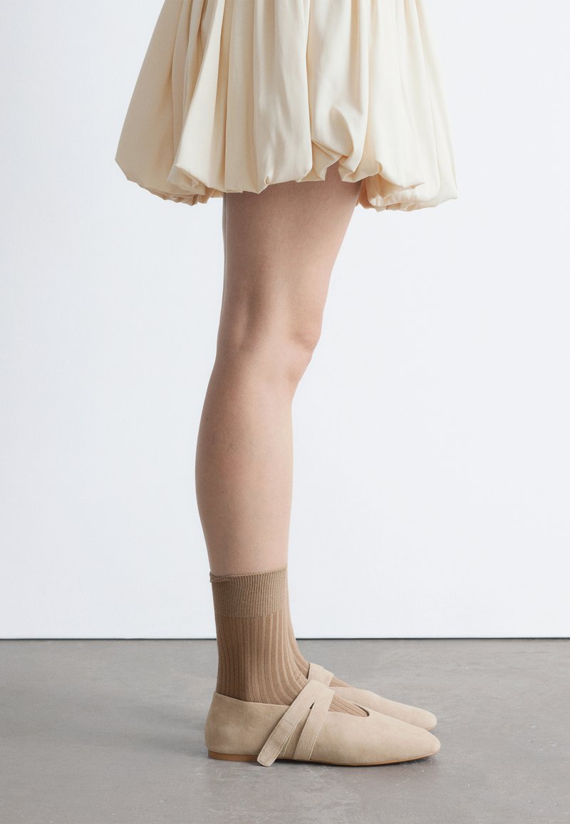Jambes portant des chaussettes côtelées beige et des chaussures plates beiges avec une bride, associées à une jupe crème bouffante sur un fond uni.