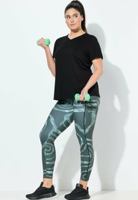 Camiseta negra de manga corta, leggings verdes con patrones de diseños en espiral, sosteniendo mancuernas verde claro, zapatillas deportivas negras.