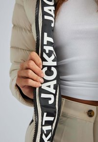 Lysebeige dunjakke med sort lynlåsbånd med fed hvid tekst "JACKIT." Nærbillede af hånd, der griber fat i lynlåsen. Tekstureret stof.