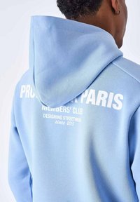 Sudadera de color azul claro, confeccionada en un tejido suave, con capucha y texto impreso en blanco en la parte posterior que dice "PROTEGER EL CLUB DE MIEMBROS DE PARÍS."