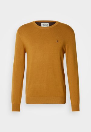 Žuta pletiva svjetlucava s okruglim dekolteom, dugim rukavima i rebrastim manžetama. Sadrži mali crni logo na prsima.