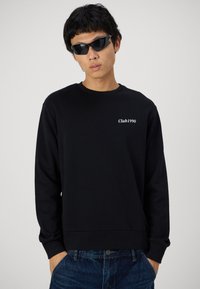 Zwarte sweatshirt met lange mouwen, geribbelde manchetten, ronde hals, en witte "Club1990" tekst op de borst, gecombineerd met donkere spijkerbroek en zonnebril.