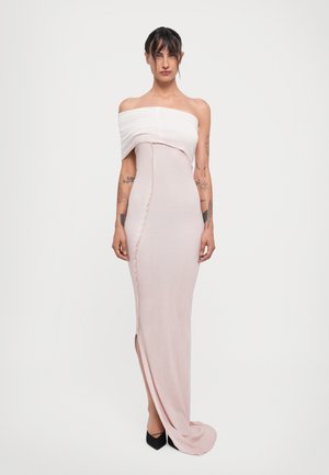 Donna con tatuaggi che indossa un abito aderente rosa pallido con spalle scoperte, dotato di uno spacco frontale, e scarpe nere con punta, su sfondo bianco.