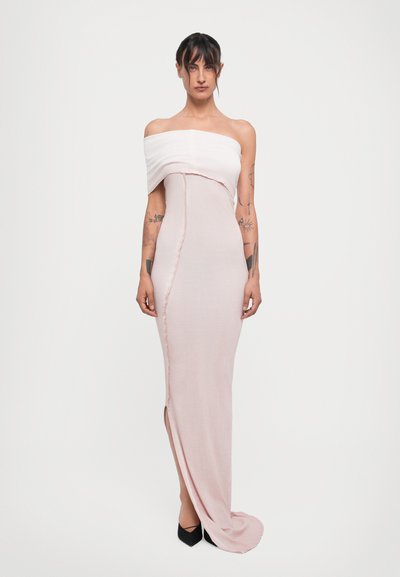 Woman with tattoos wearing a pale pink off-shoulder fitted gown with a front slit and black pointed shoes on white background.