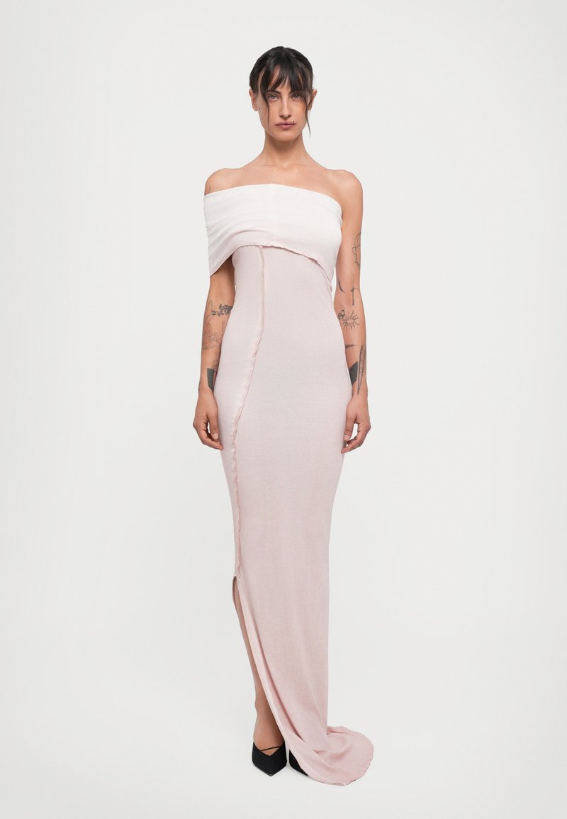 Femeie cu tatuaje îmbrăcată într-o rochie ajustată, off-shoulder, de culoare roz pal, cu o deschidere frontală și pantofi cu vârf ascuțit negri, pe un fundal alb.