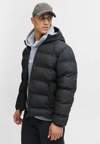 Młody mężczyzna w czarnej kurtce puchowej, nałożonej na szary hoodie i czapkę, patrzy w lewo na jednolity jasny tle.