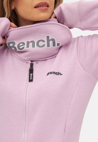 Purple zip-up hoodie with a high collar, featuring bold black "Bench." logo. Smooth fabric with a visible zipper detail at the front.