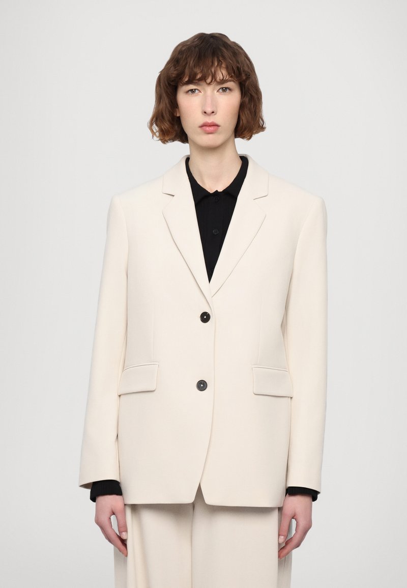 Off-white blazer z oblikovanimi rameni, z dvema sprednjima žepoma in črnimi gumbi. Nosi se čez črno srajco z gladko teksturo.