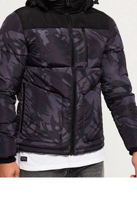 Puffjacka i lila och svart med kamouflagemönster, dragkedja fram, dragsko i huvan och sidofickor. Texturerad materialdetalj synlig.