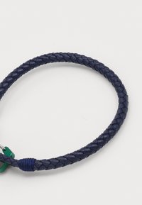Bracciale in pelle intrecciata blu navy con texture liscia e chiusura in metallo verde, caratterizzato da un piccolo filo blu accentato vicino alla chiusura.