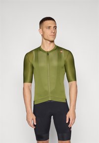 Olivgrünes Fahrradtrikot mit kurzen Ärmeln, ausgestattet mit einem Reißverschluss auf der Vorderseite und einem strukturierten Muster. Kombiniert mit schwarzen Fahrradhosen.