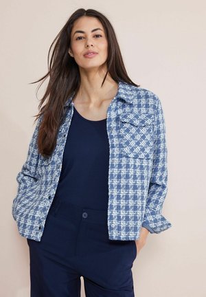 Camicia a quadri blu e bianca con maniche lunghe, chiusura a bottoni e tasche sul petto, abbinata a una canotta blu navy e pantaloni blu scuri.
