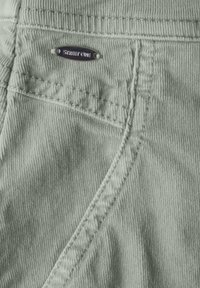 Grüner Cordstoff mit sichtbaren Rippungen, versehen mit einer genähten Tasche und einem metallischen Etikett mit "STREET ONE".