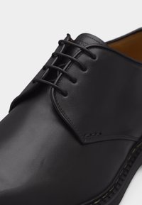 Schwarzer Schnürschuh aus Leder mit glattem Finish und sichtbaren Nähten entlang der Sohle, in Nahaufnahme gezeigt.