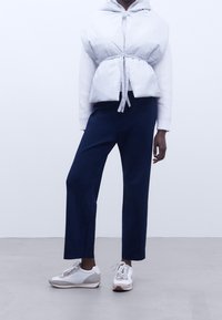 Ljusgrå ärmlös pufferväst med knytning, bärs över en vit tröja, ihop med marinblå byxor och vita sneakers med gråa accenter.