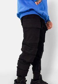 Zwarte cargobroek met meerdere opgestikte zakken en een getextureerde stof. Ontworpen met een taps toelopende pasvorm en elastische boorden. Gecombineerd met zwarte sneakers.