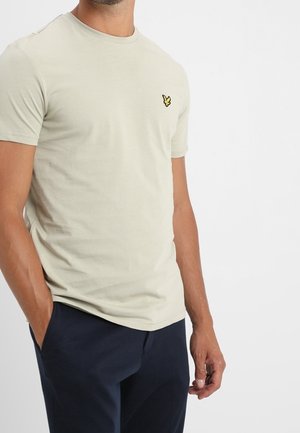 Mand iført en lysebeige kortærmet t-shirt med en lille gul fuglelogo på brystet og mørkeblå bukser, hænderne i lommerne.