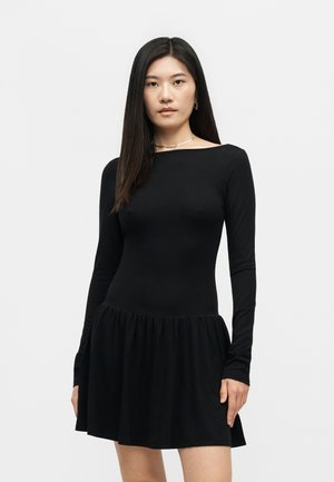 Γυναίκα με μακριά σκούρα μαλλιά που φοράει μαύρο φόρεμα με μακριά μανίκια, εφαρμοστό τοπ και φαρδιά φούστα, στέκεται σε φωτεινό φόντο.