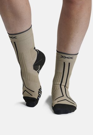 Voeten met beige en zwarte enkelcompressiekousen met geribbelde textuur en geometrische patronen, staand op een witte ondergrond.