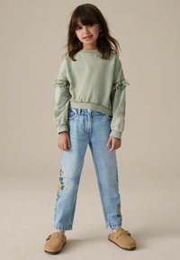 Hellgrüner Pullover mit Spitze und Rüschensäulen, kombiniert mit hellblauen, zerrissenen Jeans mit floralen Stickereien. Braune Slippers.