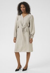 Robe beige à manches longues avec boutonnière, taille cintrée par un lien, et en tissu texturé. À porter avec des escarpins noirs à talons pointus.