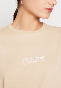 Beige bomuld T-shirt med rund hals og korte ærmer. Har hvid tekst "PACIFIC COAST California" på fronten. Glat tekstur.
