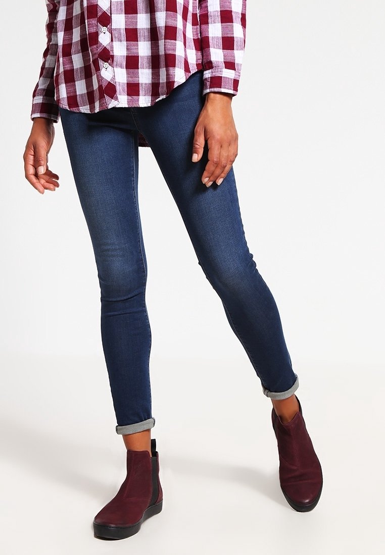 Personne portant un jean skinny bleu foncé avec les ourlets retroussés, des bottines bordeaux et une chemise à carreaux rouge et blanc, debout les jambes croisées.
