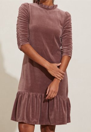 Femme portant une robe en velours mauve avec des manches trois-quarts froncées et un ourlet à volants, debout devant un fond neutre.