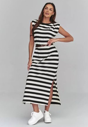 Kobieta pozuje w czarno-białej midi sukience w poziome paski, z bocznym rozcięciem, w talii z paskiem i białych trampkach na jednolitym tle.